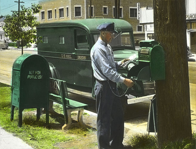 A postal worker opening a green letter box mounted to a telephone pole.