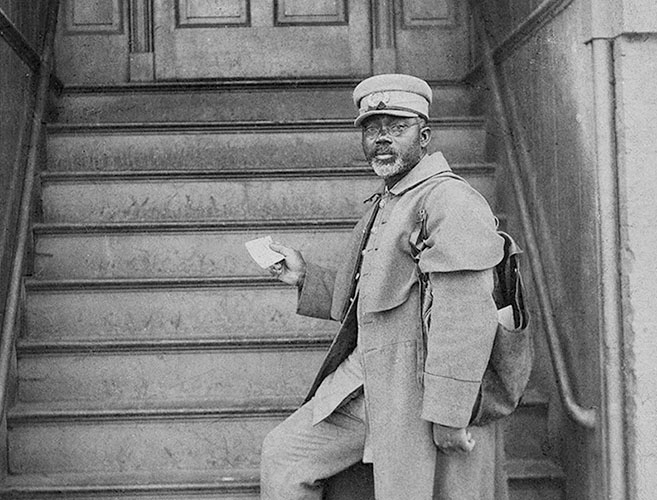 A black-and-white photograph of Sgt. William Harvey Carney holding a letter in his right hand while carrying a mail bag over his left shoulder.