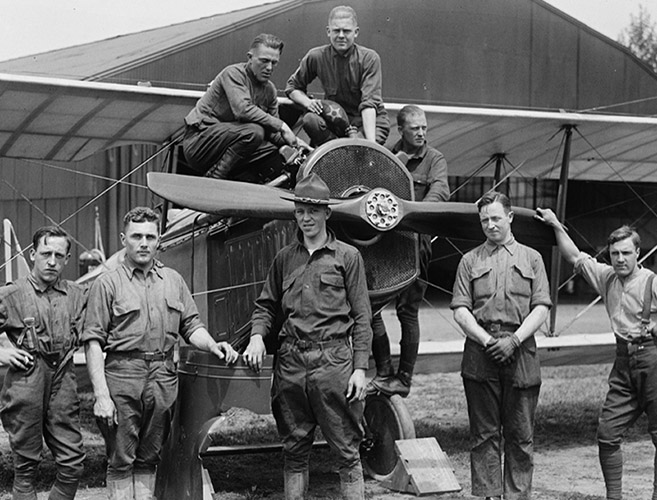 A black-and-white photograph featuring a group of men posing with a small airplane.