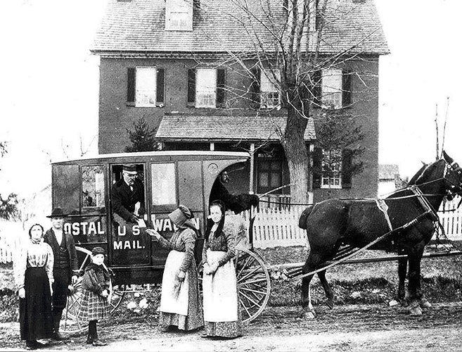 A black-and-white photograph of a family picking up mail from a postal carrier leaning out of a horse-drawn carriage.