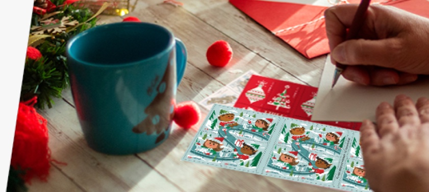 A person addressing a letter with First-Class Mail Forever® holiday stamps on the table.
