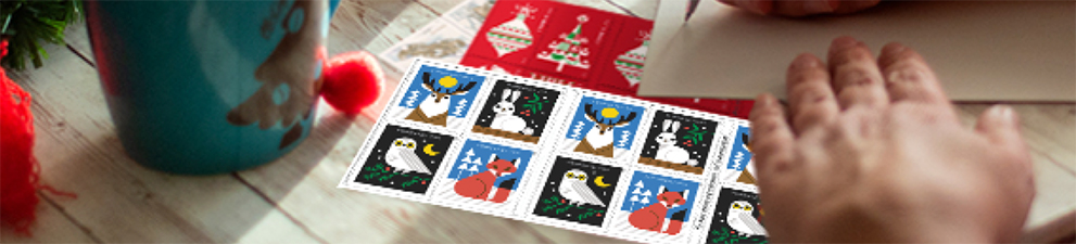 一个人正在为桌上贴着 First-Class Mail Forever® 假日邮票的一封信填写地址。