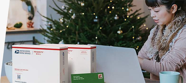 一位女士在笔记本电脑前准备运送 Priority Mail 包装盒和一张带有节日主题邮票的卡片。