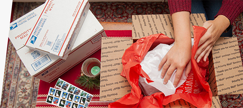 一个人正在打包节日包裹，桌子上放着 Priority Mail 包装盒和假日邮票。