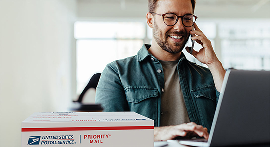 Person talking into a phone while using a laptop, with a USPS Priority Mail box in front.