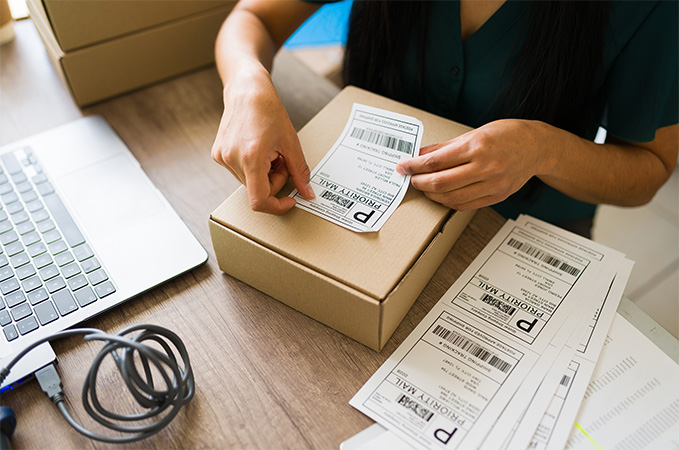 A person placing a label on a shipping box with additional labels and a laptop on the desk around them.