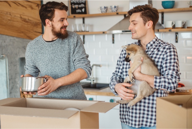 Two people packing moving boxes while holding a puppy.
