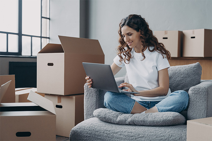 A woman sitting on couch surrounded by moving boxes changing her address online.