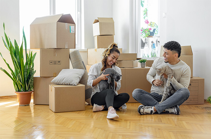 A couple sitting together with their cats surrounded by moving boxes.