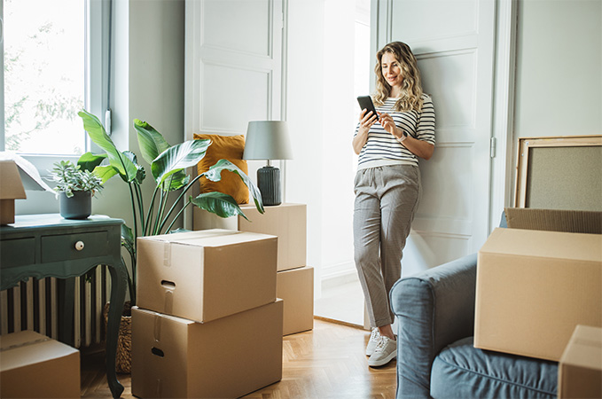 A person standing against a wall on their phone with moving boxes scattered around the room.