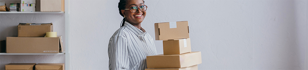 A smiling person carrying shipping boxes while a shelf behind them holds additional shipping boxes.