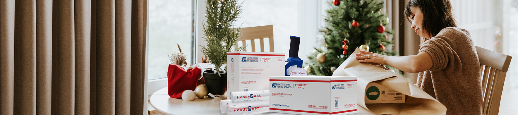 Woman wrapping holiday gifts with USPS shipping supplies on the table.