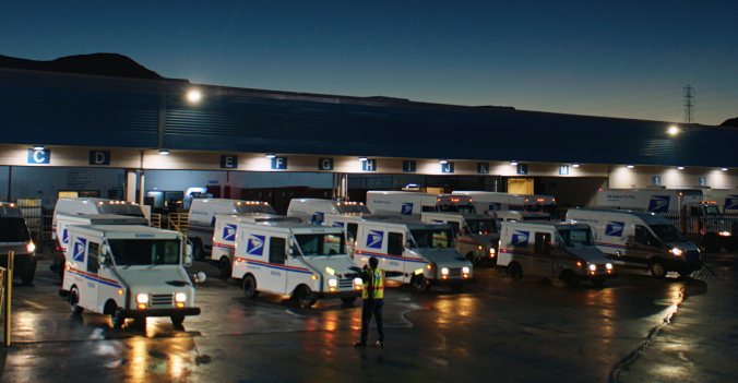 A formation of USPS delivery vehicles lines up in the early morning darkness, directed by a USPS employee in a high-visibility vest.