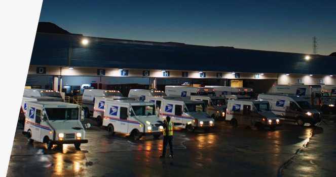 A formation of USPS delivery vehicles lines up in the early morning darkness, directed by a USPS employee in a high-visibility vest.